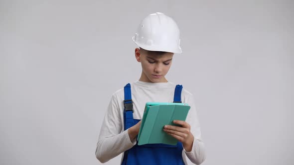 Focused Serious Boy in Hard Hat and Overalls Using Tablet Thinking and Looking at Camera alt