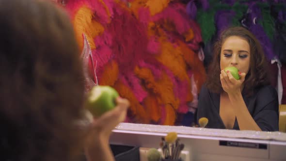 Beautiful Girl Actress with Makeup Bites Off and Eats a Green Apple in Front of the Mirror