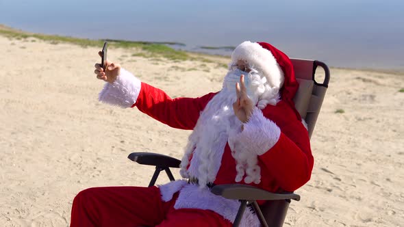 Santa Claus in a White Protective Mask Makes Selfie Lying on a Sun Lounger on the Beach alt