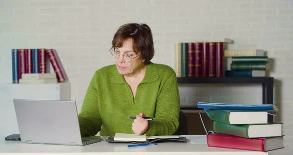 Senor Adult Woman in a Green Sweater Works in Front of a Laptop Monitor in the Evening Office