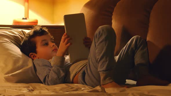 Little Boy Lays In A Bed And Touches On A Tablet Pc In The Evening