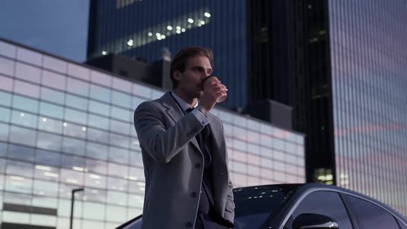 Portrait of a Cheerful Businessman Standing Near a Business Car and Drinking Coffee Top Manager in a alt
