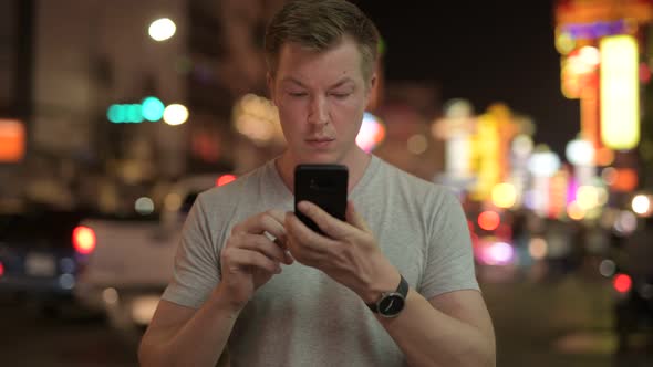 Young Happy Tourist Man Using Phone Against View of the Streets in Chinatown at Night alt
