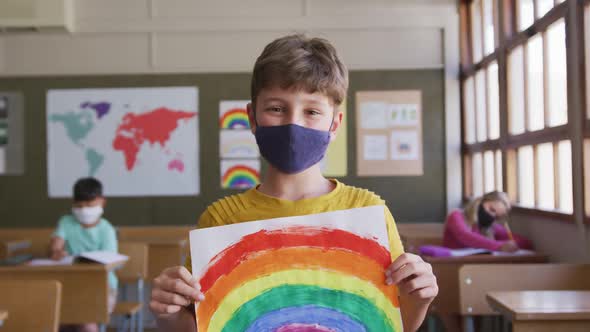 Boy wearing face mask holding a rainbow painting in class at school  alt