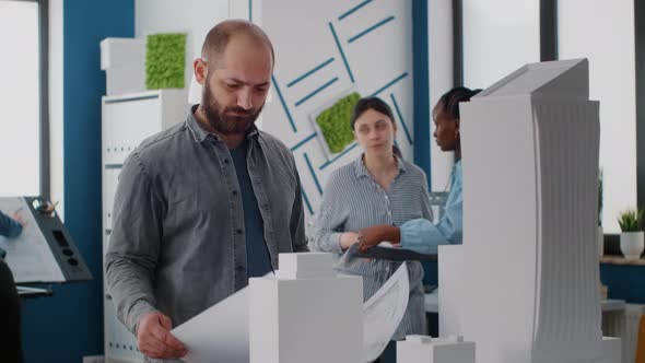 Portrait of Man Architect Looking at Blueprints Plan and Building Model on Table alt