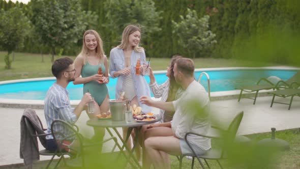 Group of happy young people cheering with drinks and eating fruits by the pool in the garden