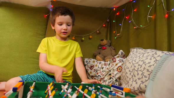 Lovely Caucasian Boy Playing Table Football in a Small House with Blankets and Pillows Is Decorated