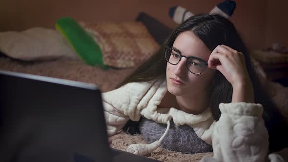 Girl student with glasses prepares homework on a laptop while lying in her bed in the evening alt