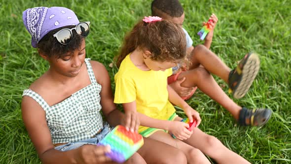 Children of Different Races Sit on the Grass and Play with an Antistress Toy Popit alt