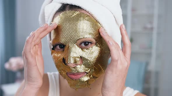 Smiling Woman Fixing Rejuvenating Cosmetic Golden Tissue Mask on Face alt