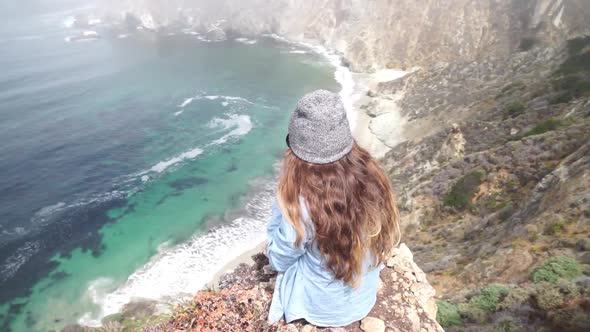 A young woman sitting on top of a cliff with the Bixby Creek Bridge in the background. alt