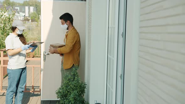 Man in Medical Mask Receiving Package From Delivery Woman Signing Paper During Covid19 Lockdown alt