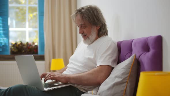 Senior Man Sitting on Bed Working Online From Home with Laptop alt