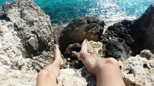 First Person Perspective of Young Man Sitting on Rock Cliff Edge Above Sea alt