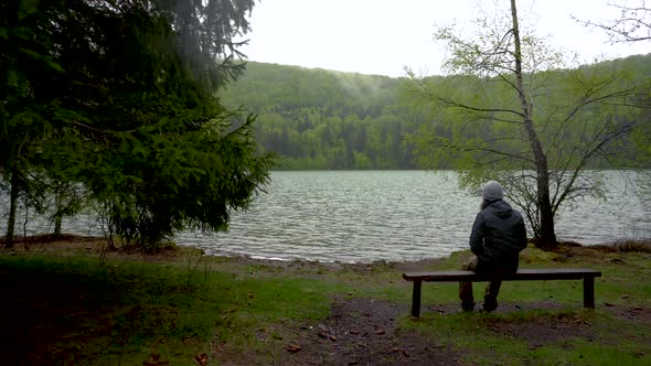 Enigmatic lonely man sitting on bench on lake shore in the mountain forest. Lake Sf. Ana Romania alt
