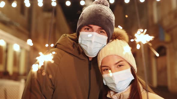 Couple in Protective Medical Masks Standing on the Street Holding Sparklers of Christmas Holidays