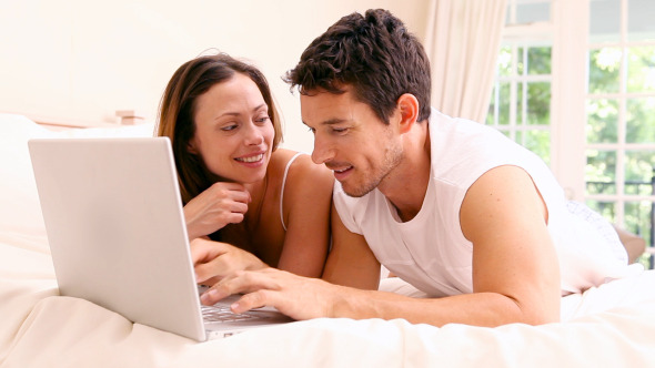 Happy Couple Lying On Bed Using Laptop Together
