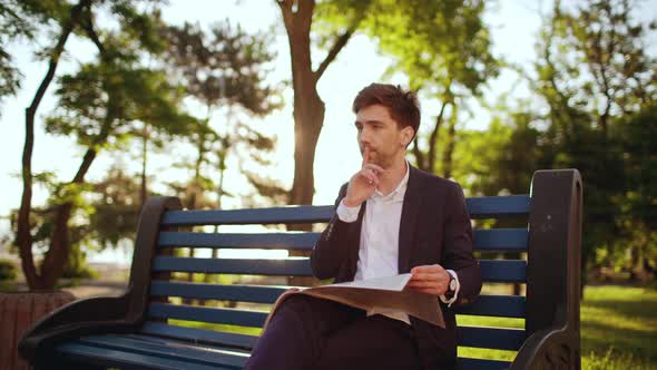 Slow Motion of Young Businessman Salesman in Suit Sitting on Bench in Green Park Reading Newspaper