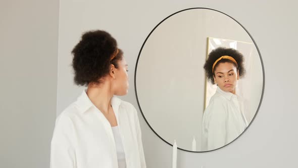 African american woman getting dressed looking in mirror. Trying on white shirt clothes alt