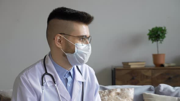 Close-up of a Young Doctor, a Man in Glasses and a Mask, a White Coat, Sitting on a Sofa at Home alt