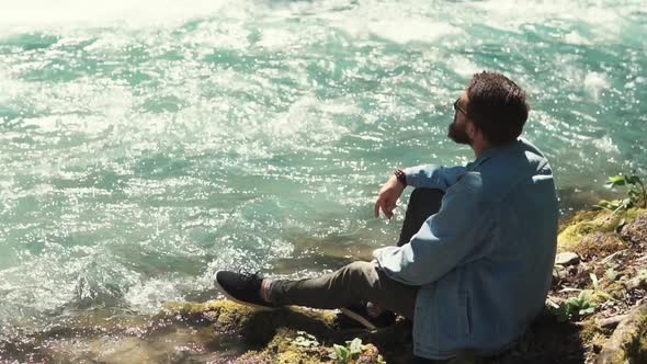 a Young Man Sits Near a Fast Stream of a River, He Enjoys the Wind and Nature alt