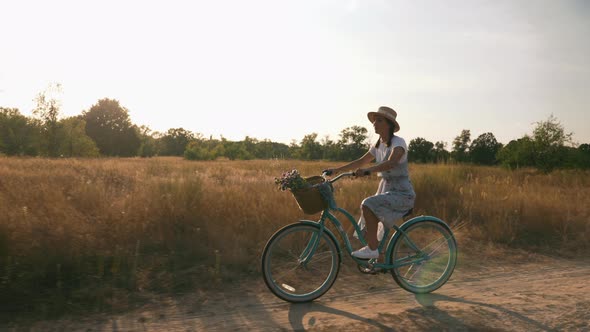 Cheerful smiling woman on retro vintage bicycle rides along sunset filled meadow.  alt