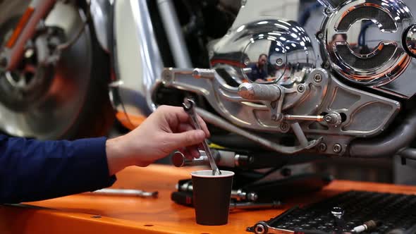 Man Stirs Coffee with a Wrench Near the Motorcycle in a Workshop