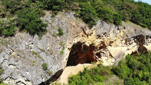 Open cave in rocky hill, all covered with green bushes on top, aerial view alt