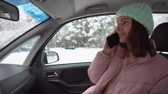 Young Beautiful Girl in a Pink Down Jacket and Winter Hat Talking Use Your Smartphone While Sitting alt
