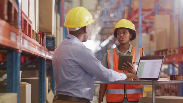 African american male and female workers wearing helmet and using laptop in warehouse alt