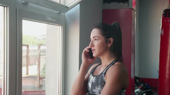 Young Woman in Sport Outfit is Standing Near the Window Talking on the Phone and Drinks Multivitamin alt