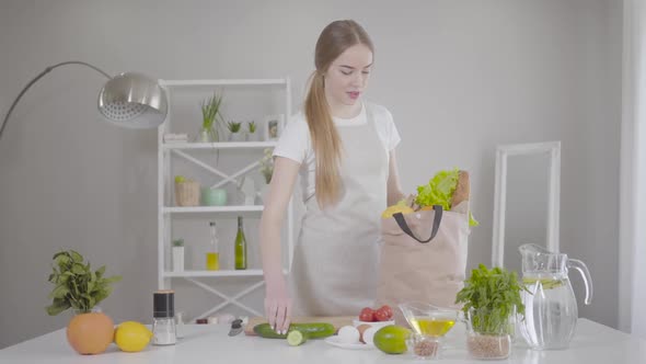 Confident Young Caucasian Woman Taking Out Fruits and Vegetables From Bag at Home alt