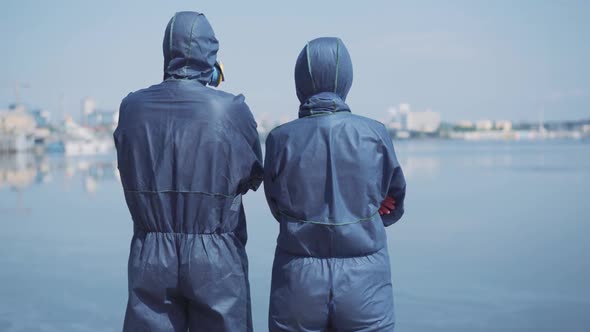 Back View of Two People in Biohazard Suits Standing at Riverbank and Looking at Blurred City on the alt