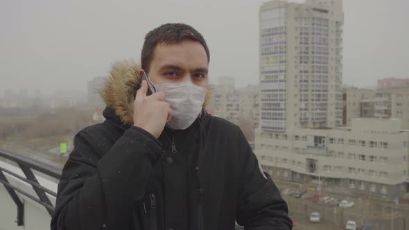 Man in a Medical Mask Speaks Seriously on the Phone Cityscape in the Background