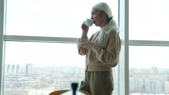 Portrait of Mature Woman Standing Near Big Window Drinking Coffee and Smiling alt