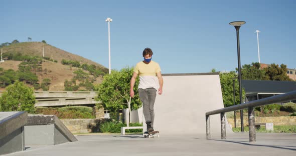 Caucasian man wearing face mask, riding and jumping on skateboard on sunny day alt