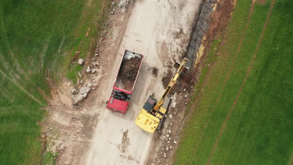 Top View Yellow Excavator Picks Up Land From the Field and Loads It Onto a Red Truck alt