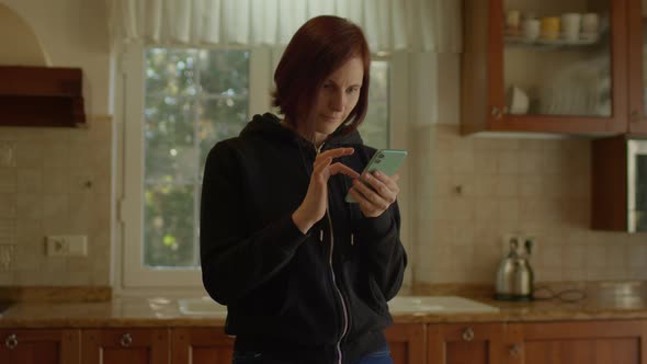 30s Woman Reads Good News on Mobile Phone Standing in the Kitchen alt
