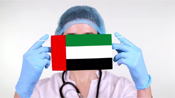 Close-up. Doctor in Glasses, Blue Medical Cap, Gloves Holds in Hands Medical Mask with UAE Flag alt