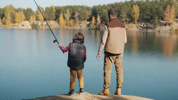 Back View of Adult Man and Little Boy Fishing in Lake on Sunny Autumn Day alt