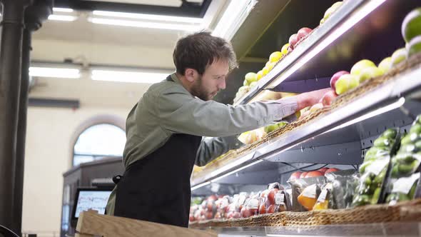 Busy Salesman is Putting Apples on Shelves at Fruits Section in Supermarket Bearded Guy is Wearing alt