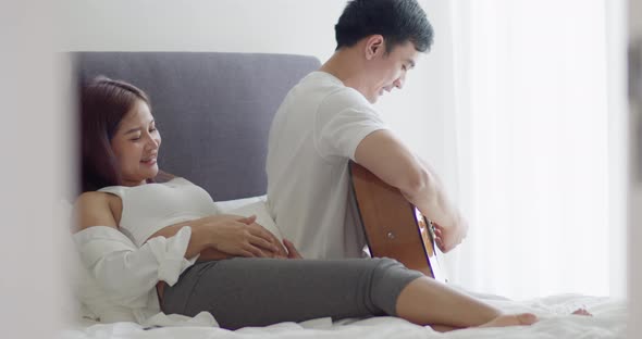 Asian husband and pregnant wife spend time together in the bedroom and husband play guitar.