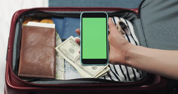 Young Woman Getting Ready for a Trip. She Holding Smartphone with Green Screen on the Suitcase alt
