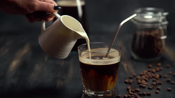 Slow Motion of Cream Being Poured Into a Glass of Coffee on Black Wood Table with Metal Cocktail