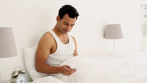 Cheerful Handsome Man Sitting On Bed Texting