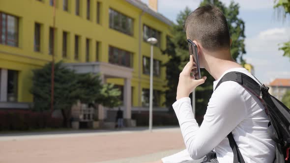 A Caucasian Teenage Boy Talks on a Smartphone  Side View  a School in the Background