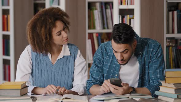 Two Diverse Students Sitting in Library Preparing for Exam Young Guy Enthusiastically Plays on Phone alt