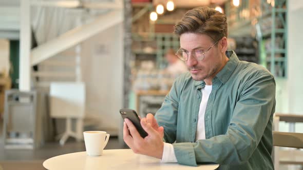 Middle Aged Man Celebrating Success on Smartphone in Cafe alt