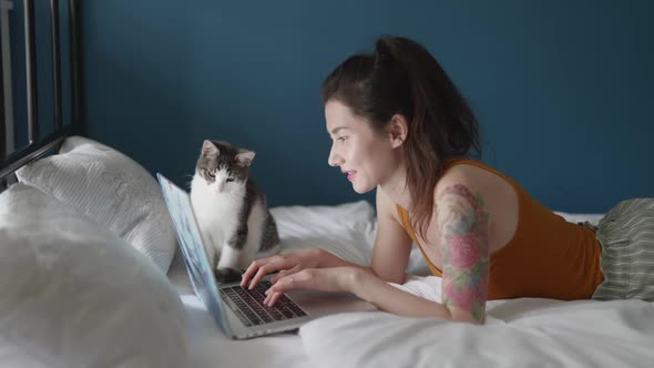 Young Asian Woman Working on Laptop Online at Home Lying on Bed in Bedroom with Her Cat alt