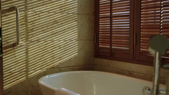 white bathtub in clean bathroom with big window and wooden blindshade with light evening time